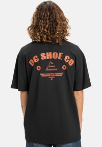 Zwarte katoenen t-shirt met een grote oranje grafische tekst op de rug, waarop staat "DC SHOE CO," met sterren en extra tekstdetails eronder.