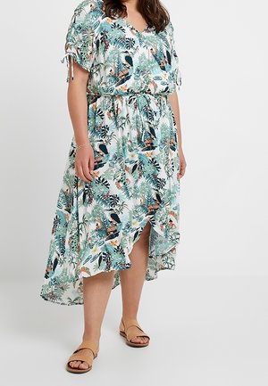 Kobieta w midi sukience z paskiem, krótkim rękawem i dekoltem w kształcie litery V, ozdobionej nadrukiem tropikalnych ptaków i liści, w połączeniu z brązowymi płaskimi sandałami.