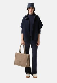 Navy gestrickter Poncho mit Kapuze, kombiniert mit einem figurbetonten Oberteil und weit geschnittenen Hosen. Beige Tote-Bag mit strukturiertem Muster und Logo. Weiße, flauschige Schuhe.