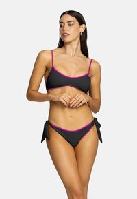 Bikini nero con dettagli fucsia, caratterizzato da un top a halter e un fondo con lacci laterali. Texture liscia con dettagli in hardware minimi.