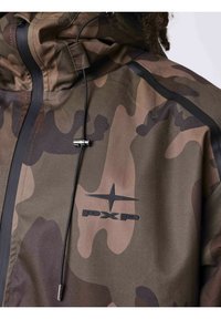 Gros plan d'une personne portant une veste camouflage avec une fermeture éclair, un cordon noir et un logo PXP sur la poitrine.