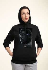 Sudadera negra con un estampado frontal que presenta un retrato monocromático de un hombre. Puños acanalados, capucha con cordón y una textura de tela suave.