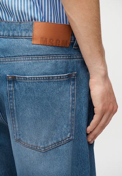 Gros plan sur une personne portant un jean en denim bleu avec un patch en cuir marron et une chemise rayée bleue rentrée à l'intérieur.