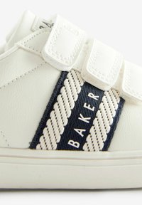 Zapatilla de cuero blanca con dos correas de velcro, que presenta una franja de tela azul marino decorada con un patrón trenzado y el texto "BAKER."