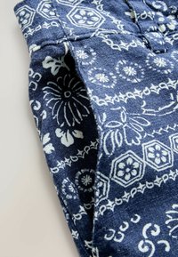 Tissu bleu avec motifs floraux et géométriques blancs, montrant une section pliée et un détail de couture ou de poche.