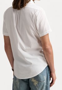 Kortärmad vit skjorta i texturerad vadmal med rundad nederkant. Har en klassisk krage och en veckdetalj bak för ökad rörelsefrihet.