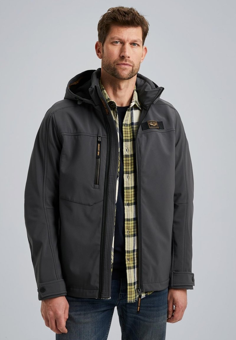 Gray jacket with a hood, featuring multiple zippered pockets and a textured surface. Worn over a plaid shirt, paired with blue jeans.