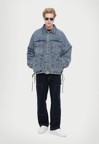 Jachetă din denim supradimensionată cu două buzunare frontale, de culoare albastră, cu tiv ajustabil, asortată cu pantaloni negri cu cusături contrastante și adidași albi.