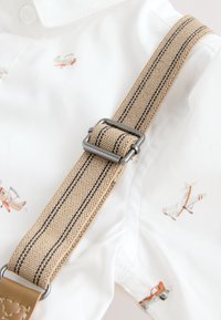 Beige fabric suspenders with a metallic clip, featuring black stripes, attached to a white shirt with small airplane patterns.