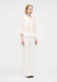 Mujer con cabello rojo hasta los hombros, vistiendo una blusa blanca transparente con mangas con volantes, pantalones blancos y zapatos oscuros de punta, de pie sobre un fondo liso.