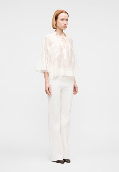 Γυναίκα με κόκκινα μαλλιά μέχρι τους ώμους που φορά ημιδιάφανο λευκό πουκάμισο με βολάν στα μανίκια, λευκό παντελόνι και σκούρα μυτερά παπούτσια, στέκεται σε απλό φόντο.