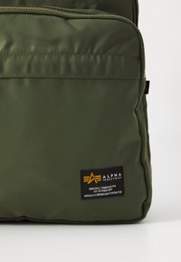 Mochila de nylon verde oliva con una textura suave, con una cremallera dorada y una etiqueta negra que muestra el nombre de la marca y los detalles.