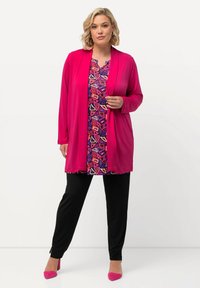 Helles pinkes Cardigan über einem mehrfarbig gemusterten Oberteil mit geometrischen Formen, kombiniert mit schwarzen Hosen und pinken Absatzschuhen.