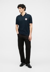 Νέος άνδρας που στέκεται με τα χέρια στις τσέπες, φορώντας μπλε σκούρο πόλο με έμβλημα και μαύρο παντελόνι και παπούτσια, κοιτάζοντας πλάγια σε απλό φόντο.