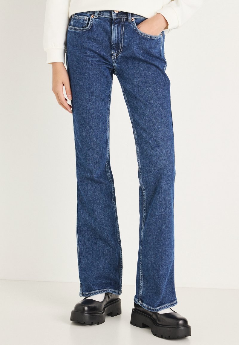 Personne portant un jean bleu droit avec une main dans la poche, associé à des chaussures noires épaisses et des chaussettes blanches sur un fond uni.