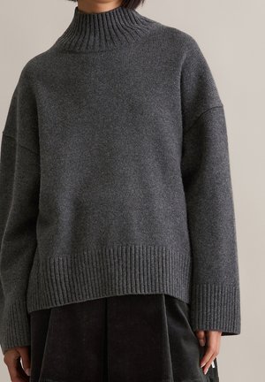 Person iført en mørkegrå, ribstrikket rullekravetrøje med lange ærmer og mørke bukser foran en ensfarvet baggrund.