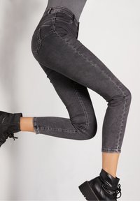 Piernas en jeans skinny negros desgastados y botines negros con cordones, con una rodilla doblada hacia arriba contra un fondo neutral.