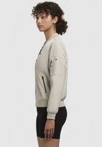 Jachetă bomber bej cu închidere cu fermoar, manșete ribbate și un buzunar pe braț. Material neted cu o siluetă curată. Purtată peste pantaloni scurți negri.