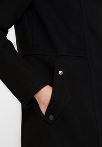 Abrigo de lana negro con una superficie texturizada, bolsillo inclinado con botón de metal plateado y detalle de manga acanalada.