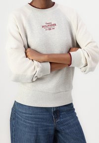 Gray sweatshirt with ribbed cuffs and hem, features embroidered red logo "Tommy Hilfiger Est. 1985," and contrasting cream sleeves.