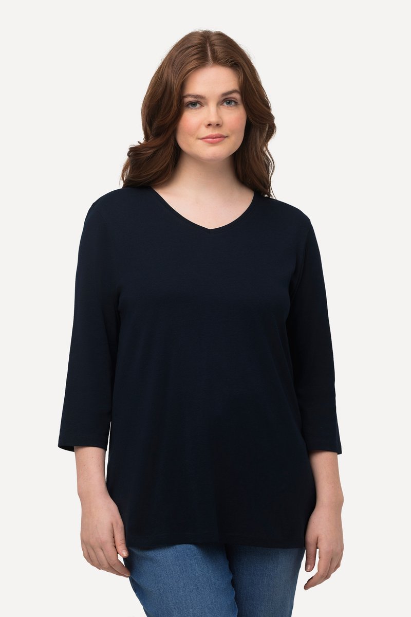 Vrouw met golvend bruin haar die een marineblauw V-hals top met driekwart mouwen en blauwe spijkerbroek draagt, staand tegen een effen lichte achtergrond.