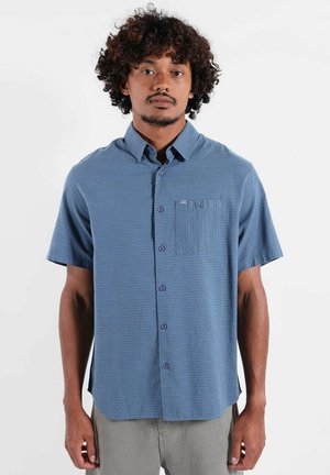 Hombre con cabello rizado que lleva una camisa de manga corta azul con textura y botones, y pantalones grises, de pie contra un fondo blanco liso.