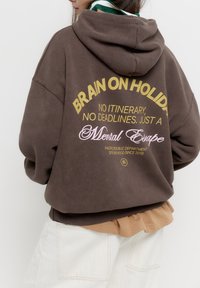 Hnědá oversized mikina s grafickým textem na zádech v žluté a růžové barvě. Obsahuje kapuci na šňůrku a žebrované manžety. Nositá přes béžové tričko.