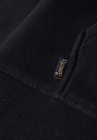 Gros plan sur un tissu noir à texture côtelée avec une petite étiquette marine portant l'inscription "Superdry" en lettres dorées.
