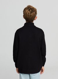 Czarny sweter z dzianiny o wysokim kołnierzu i teksturowanymi rękawami w splocie warkoczowym, zestawiony z jasnoniebieskimi dżinsami, widziany od tyłu.