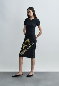 Femeie în picioare, purtând o rochie neagră mulată cu logo-ul auriu „EA7 Emporio Armani”, încălțată cu pantofi slingback negri, pe un fundal gri simplu.