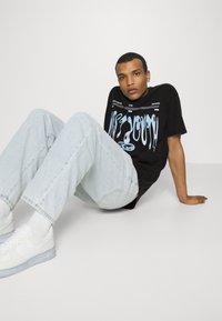  camiseta gráfica negra con diseño azul, jeans azul claro y zapatillas blancas. Posición sentado contra un fondo liso.