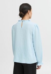Blusa azzurro chiaro a maniche lunghe con tessuto testurizzato, colletto arricciato, apertura a bottone sul retro e polsini arricciati. Pantaloni neri visibili.