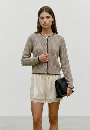 Femme portant un cardigan beige en tricot et un short en dentelle crème tenant une pochette noire sur un fond clair uni.