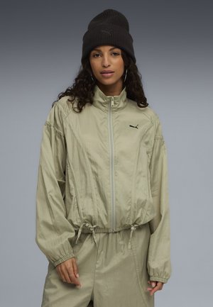 Light olive green zip-up jacket with a high collar and adjustable drawstring waist, paired with a black knit beanie. Simple, functional design.