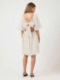 Weißes Kleid mit einem strukturierten Blumenmuster, kurzen Puffärmeln, offenem Rücken mit einer Schleife und gerafftem Bund. Schwarze Riemchenabsätze.