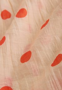 Vaalean persikanvärinen kangas, jossa on rypytetty rakenne ja hajanaisesti suuria punaisia pilkkuja.