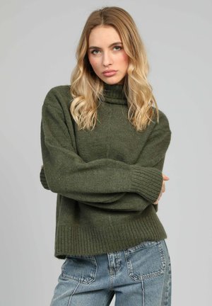 Jeune femme aux cheveux blonds portant un pull à col roulé vert et un jean bleu, debout les bras croisés devant un fond gris.