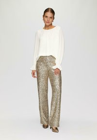 Weiße Bluse mit gerafftem Ausschnitt und langen, rüschigen Ärmeln, kombiniert mit goldenen, glitzernden weit geschnittenen Hosen. Braune hochhackige Schuhe.