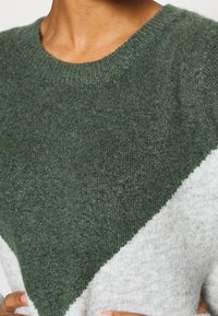 Maglione lavorato a maglia con un design a blocchi di colore triangolari, composto da sezioni di verde scuro e grigio chiaro. Texture morbida con scollatura rotonda.
