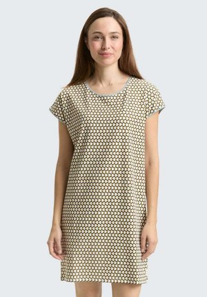 Vrouw met lang bruin haar, die een korte mouwen jurk draagt met een geometrisch patroon in wit, bruin en lichtblauw tegen een effen achtergrond.