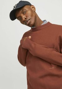 Suéter marrón de punto con cuello redondo acanalado y agujeros para los pulgares, superpuesto a una camisa a rayas azul y blanca, combinado con una gorra azul marino.