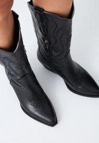 Botas de vaquero de cuero negro con puntas afiladas, cremalleras visibles y patrones florales grabados en el caña y el pie. Textura suave.