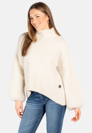 Smilende kvinne iført overdimensjonert hvit turtleneck-genser med ballongerm og blå jeans, stående mot ensfarget bakgrunn.