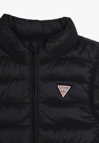 Czarna pikowana kamizelka z wysokim kołnierzem, zapinana na zamek, z trójkątną naszywką z logo. Posiada pikowaną fakturę i jednolite czarne boki.