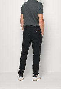 Schwarze Hosen mit glatter Oberfläche und elastischen Bündchen, kombiniert mit einem dunkelgrauen Kurzarmhemd. Auffälliges Design der Gesäßtasche.