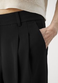 Schwarze Hose mit tailliertem Schnitt, ausgestattet mit Messerfalten und einer Seitentasche. Hergestellt aus einem glatten Stoff mit leichtem Glanz.