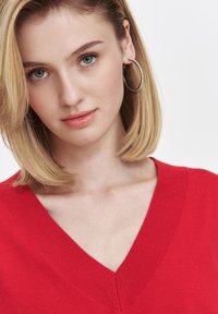 Czerwony sweter z dekoltem w serek i żebrową teksturą. Modelka ma włosy do ramion w kolorze blond i srebrne okrągłe kolczyki, na neutralnym tle.
