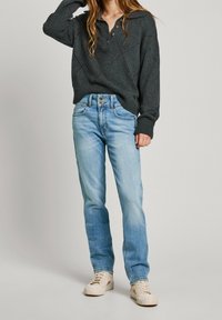 Mörkgrå stickad tröja med ett diamantsmönster och tre knappar, kombinerad med ljusblå raka jeans och krämfärgade sneakers.