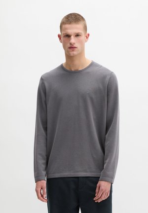 Junger Mann mit kurzen Haaren, der ein schlichtes graues Langarmshirt und dunkle Hosen trägt, stehend vor weißem Hintergrund.