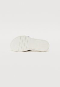 Semelle de chaussure blanche avec un motif texturé et des rainures, de forme incurvée, présentant un logo en relief. Matériau lisse, éléments de design minimalistes.